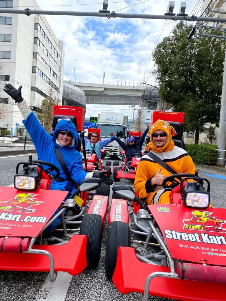 IMG_2837 - StreetKart People in red Street Kart go-karts on a city street, two in front wearing blue and orange mascot outfits, waving at the camera.