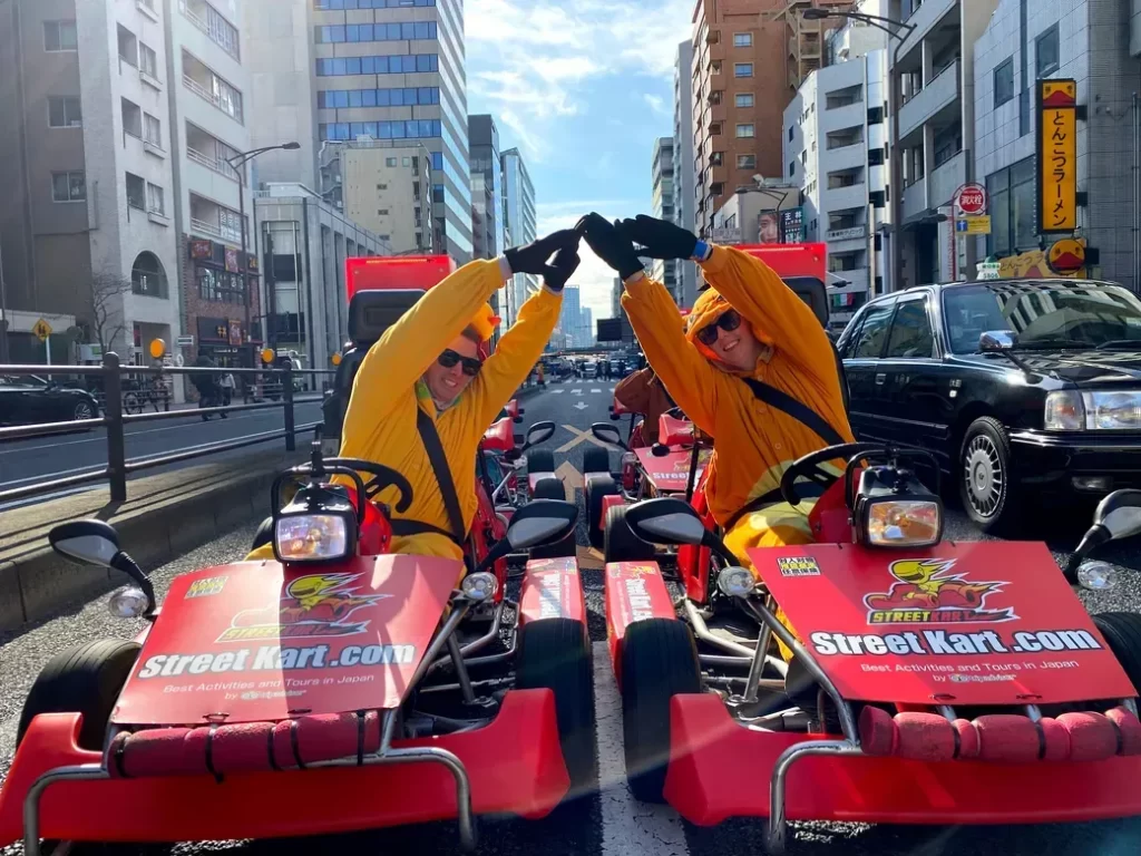 Two people in yellow outfits high-five while seated on red street-kart go-karts in an urban street scene.